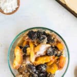 A clear glass bowl filled with mixed fruits, including peaches and blackberries, topped with a sprinkling of sugar. A small bowl of powdered sugar and a rolled-out pie crust are nearby on the countertop.