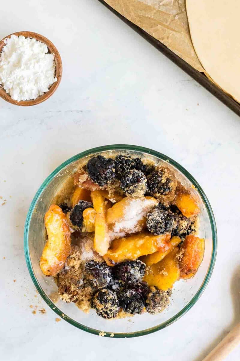 A clear glass bowl filled with mixed fruits, including peaches and blackberries, topped with a sprinkling of sugar. A small bowl of powdered sugar and a rolled-out pie crust are nearby on the countertop.