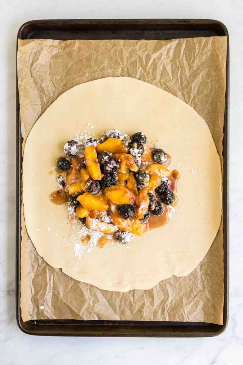 Pastry dough on a baking sheet topped with a mixture of peach slices and blueberries, prepared for baking.
