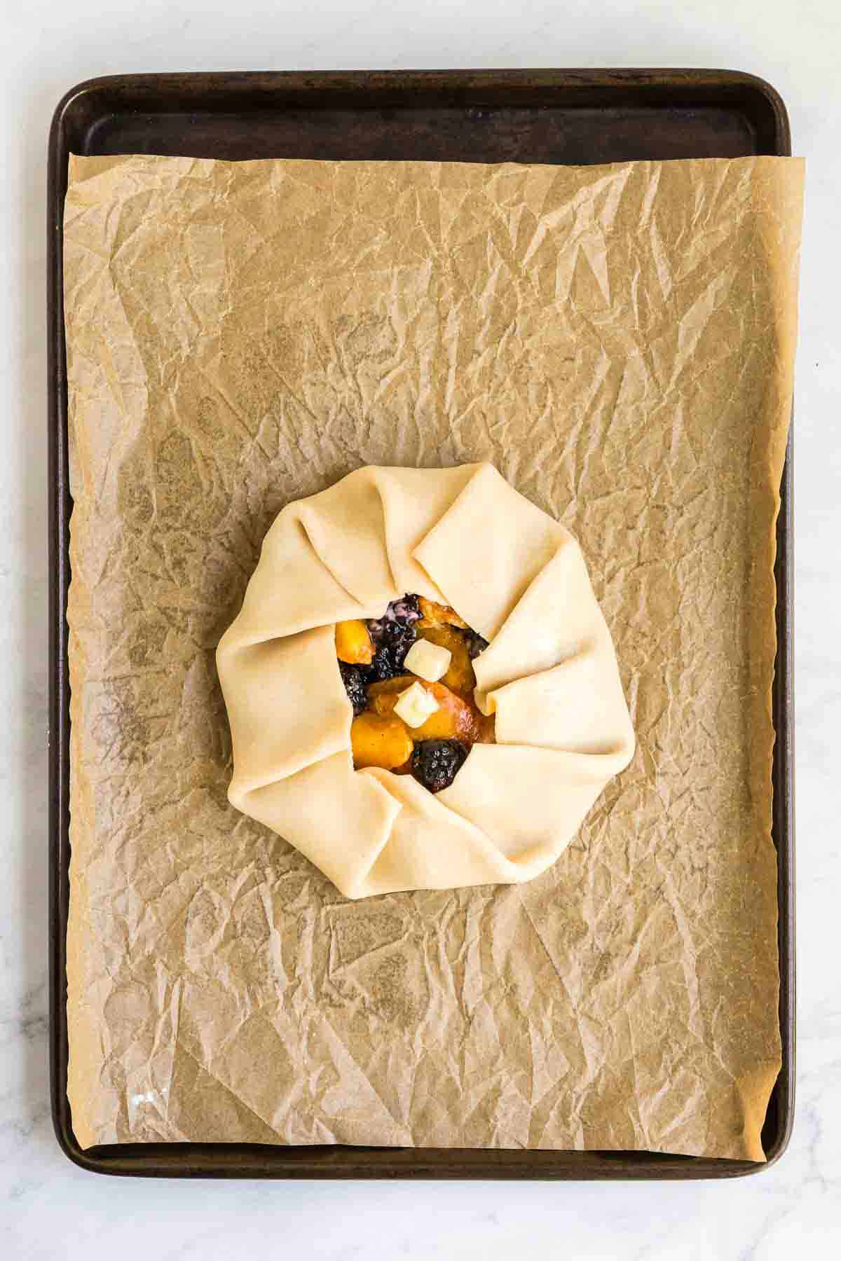 An unbaked rustic tart filled with fruit is placed on a sheet pan lined with brown parchment paper.