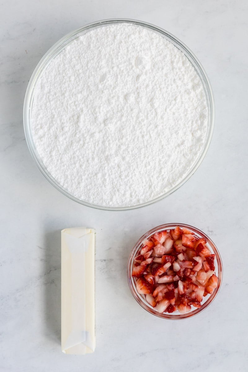 A bowl of powdered sugar, a stick of butter, and a small bowl of chopped strawberries on a light-colored surface.