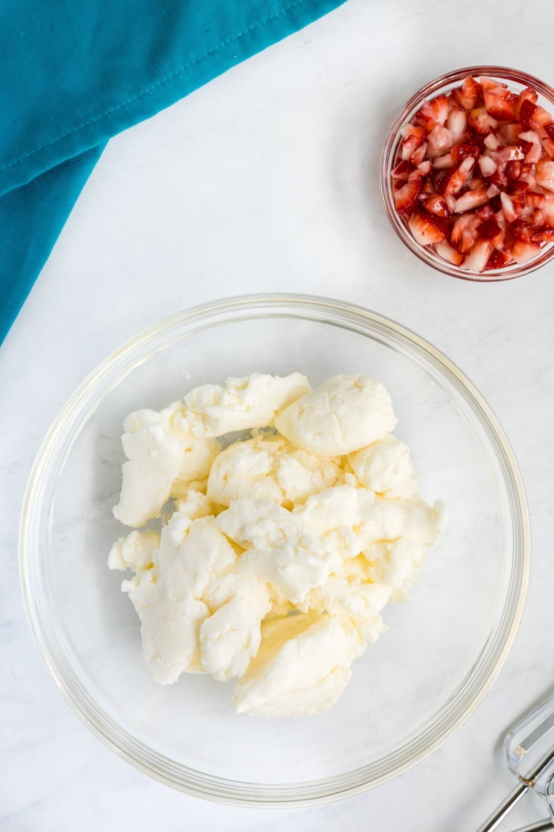 A clear glass bowl filled with butter is placed on a white surface next to a smaller bowl containing chopped strawberries. A teal napkin is partially visible in the background.