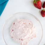 A clear bowl of strawberry buttercream frosting is on a white surface with a folded teal cloth and whole strawberries beside it.