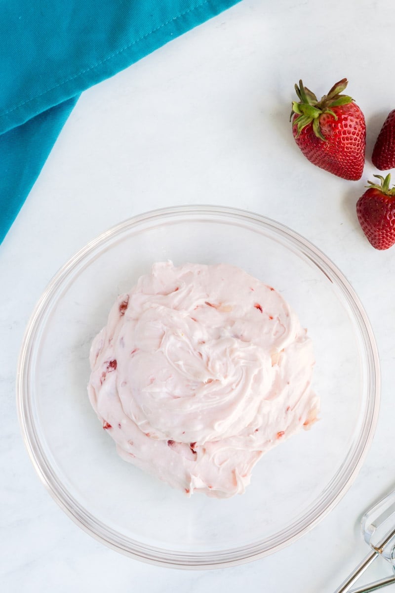 A clear bowl of strawberry buttercream frosting is on a white surface with a folded teal cloth and whole strawberries beside it.