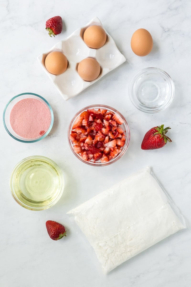 Ingredients for a strawberry jello cake are arranged on a marble surface, including eggs, sugar, diced strawberries, oil, water, flour, strawberry powder, and whole strawberries.