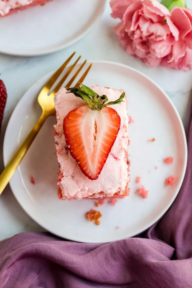 A slice of pink cake topped with a halved strawberry on a white plate, accompanied by a gold fork and a pink flower. A purple cloth is partially visible.