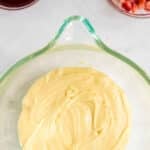 A glass bowl with yellow cake batter, a bowl of red liquid, and a bowl of diced strawberries on a white surface.