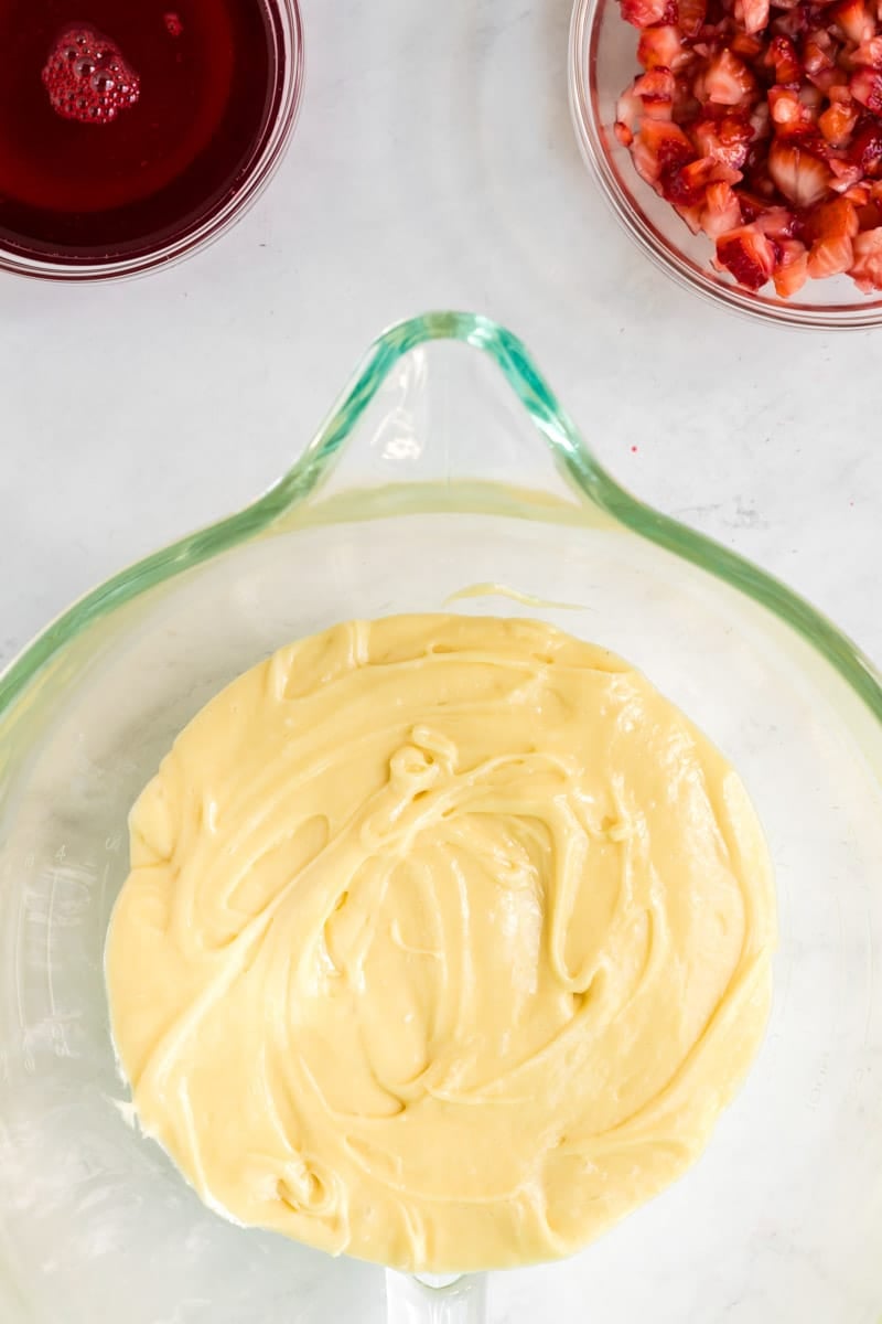 A glass bowl with yellow cake batter, a bowl of red liquid, and a bowl of diced strawberries on a white surface.