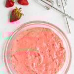 A glass bowl filled with pink strawberry batter sits on a white surface next to three whole strawberries and two detached electric mixer beaters.