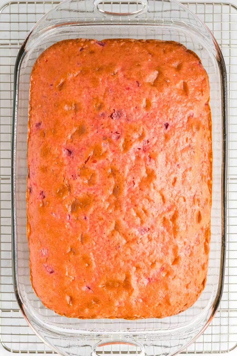 A rectangular pink cake in a glass baking dish, sitting on a wire cooling rack.