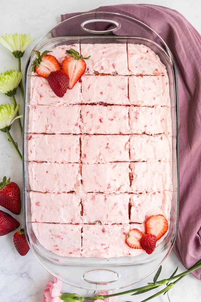 Rectangular glass dish with pink frosted cake cut into 20 square pieces, garnished with sliced strawberries. A purple cloth and fresh flowers are placed nearby on a marble surface.