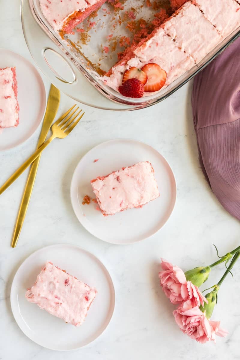 A baking dish with frosted pink strawberry cake with jello pieces, a single pink carnation, two plates with cake, a fork, a knife, and a pink cloth on a marble surface.