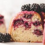A slice of blackberry cake with chunks of blackberries inside, topped with a pink glaze and fresh blackberries.