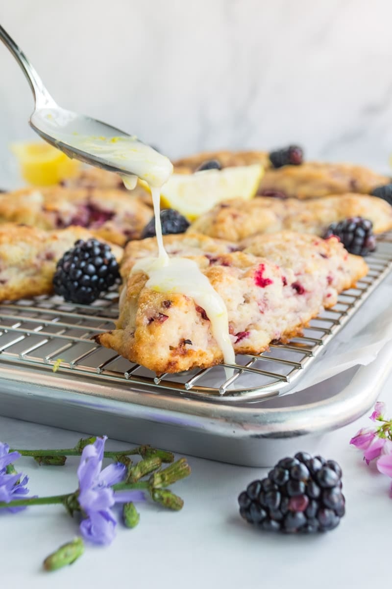 A spoon drizzles glaze over freshly baked blackberry scones on a cooling rack, with blackberries and flowers placed around.
