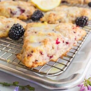 A glazed blackberry scone on a cooling rack surrounded by fresh blackberries.