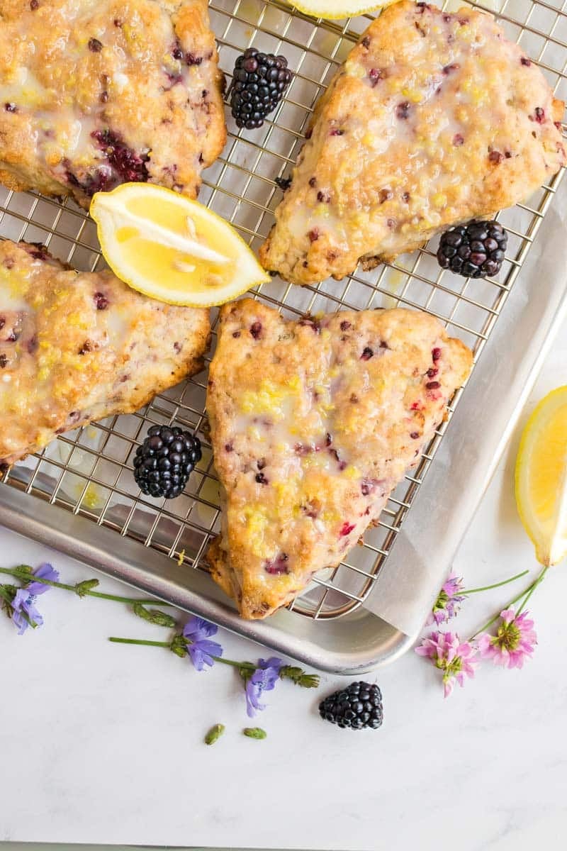 Four blackberry lemon scones with glaze on a cooling rack, garnished with fresh blackberries, lemon wedges, and small purple flowers.