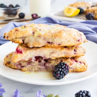 Three blackberry scones stacked on a white plate, topped with a lemon glaze. A fresh blackberry and flowers are placed around the plate. A cooling rack with additional scones is visible in the background.