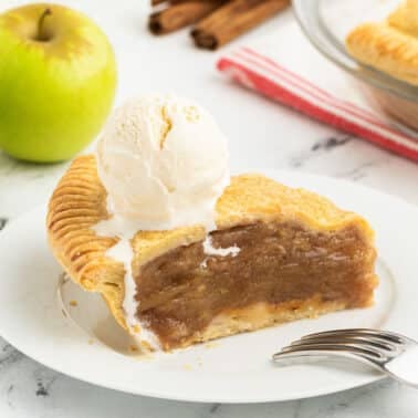 Slice of double crust apple pie topped with a scoop of vanilla ice cream on a white plate, accompanied by a stainless steel fork. Surrounding the plate are green apples, a pie dish, and cinnamon sticks on a marble surface.