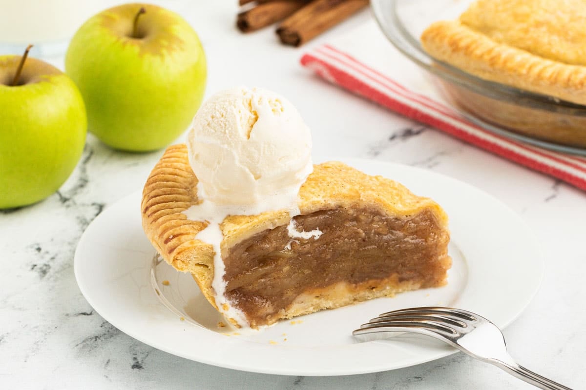 Slice of double crust apple pie topped with a scoop of vanilla ice cream on a white plate, accompanied by a stainless steel fork. Surrounding the plate are green apples, a pie dish, and cinnamon sticks on a marble surface.