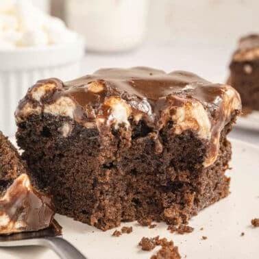 A piece of chocolate cake topped with marshmallow and frosting on a white plate, with a bite taken out and placed on a fork beside it. A bowl of marshmallows is in the background.