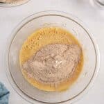 A mixing bowl containing a yellow liquid and brown powder sits on a counter, with additional bowls of ingredients nearby. A blue towel lies beside the bowl.