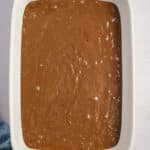 A white baking dish filled with chocolate batter on a light surface, with a small bowl of marshmallows and a small bowl of a white ingredient nearby.