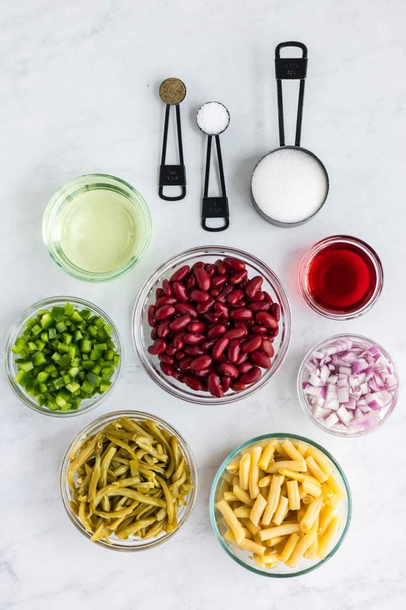 Above view of ingredients on a marble surface, including kidney beans, green beans, yellow beans, diced green bell pepper, chopped red onion, red vinegar, oil, sugar, salt, and pepper.