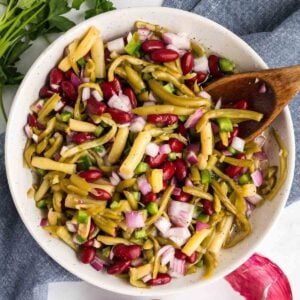 A bowl filled with a colorful three-bean salad, featuring green beans, wax beans, red kidney beans, diced red onions, and green bell peppers, mixed with a wooden spoon. Parsley garnish on the side.
