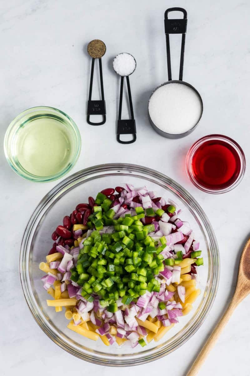 Ingredients for a bean and pasta salad are arranged on a countertop, including chopped bell pepper, red onion, macaroni, kidney beans, vegetable oil, vinegar, sugar, salt, pepper, and a wooden spoon.
