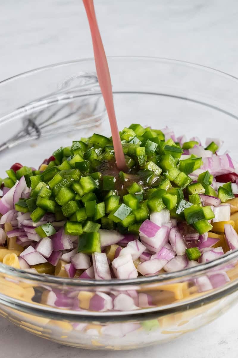 Close-up of a glass bowl containing chopped red onions, green bell peppers, and pasta, with a pink liquid being poured over the ingredients. A whisk is visible in the background on a white surface.