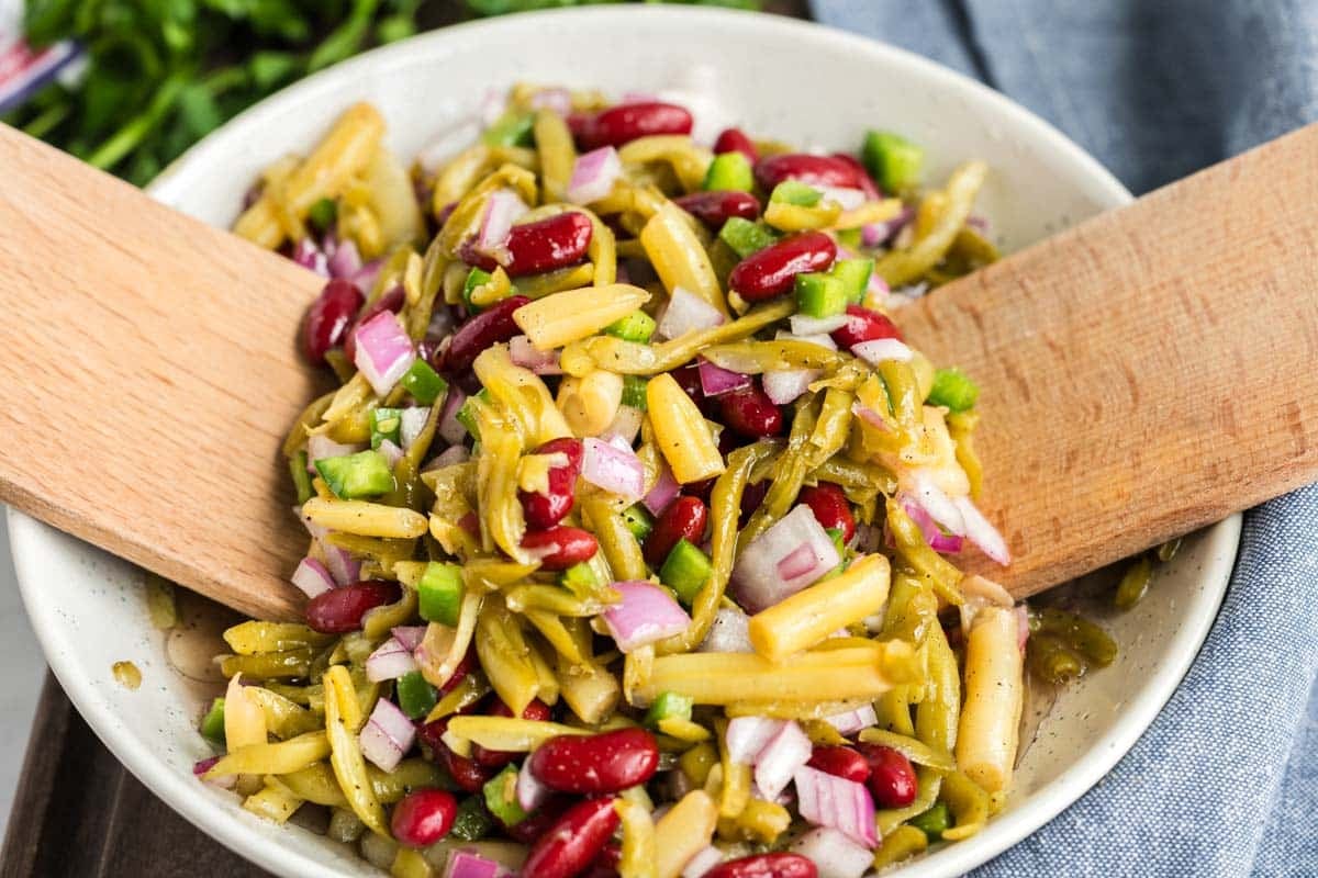 A bowl of mixed bean salad with kidney beans, green beans, wax beans, diced onions, and diced green peppers, being stirred with two wooden utensils.
