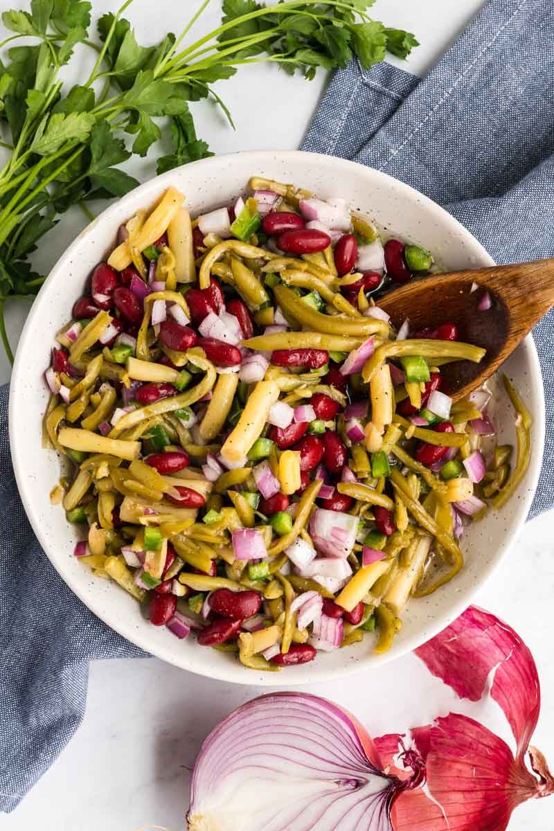 A bowl of easy three bean salad with green beans, red beans, chopped onions, and diced vegetables, garnished with parsley and placed on a grey cloth. A wooden spoon is in the bowl, with red onions nearby.
