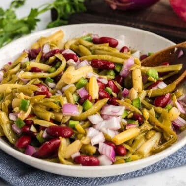 A bowl of three bean salad with chopped red onions and green vegetables, featuring kidney beans and other beans. A wooden spoon is placed in the salad.