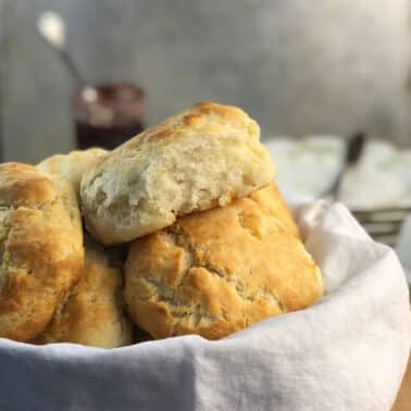 A basket filled with freshly baked biscuits, one biscuit placed partially on top of others. A jar of jam and a spoon are blurred in the background.