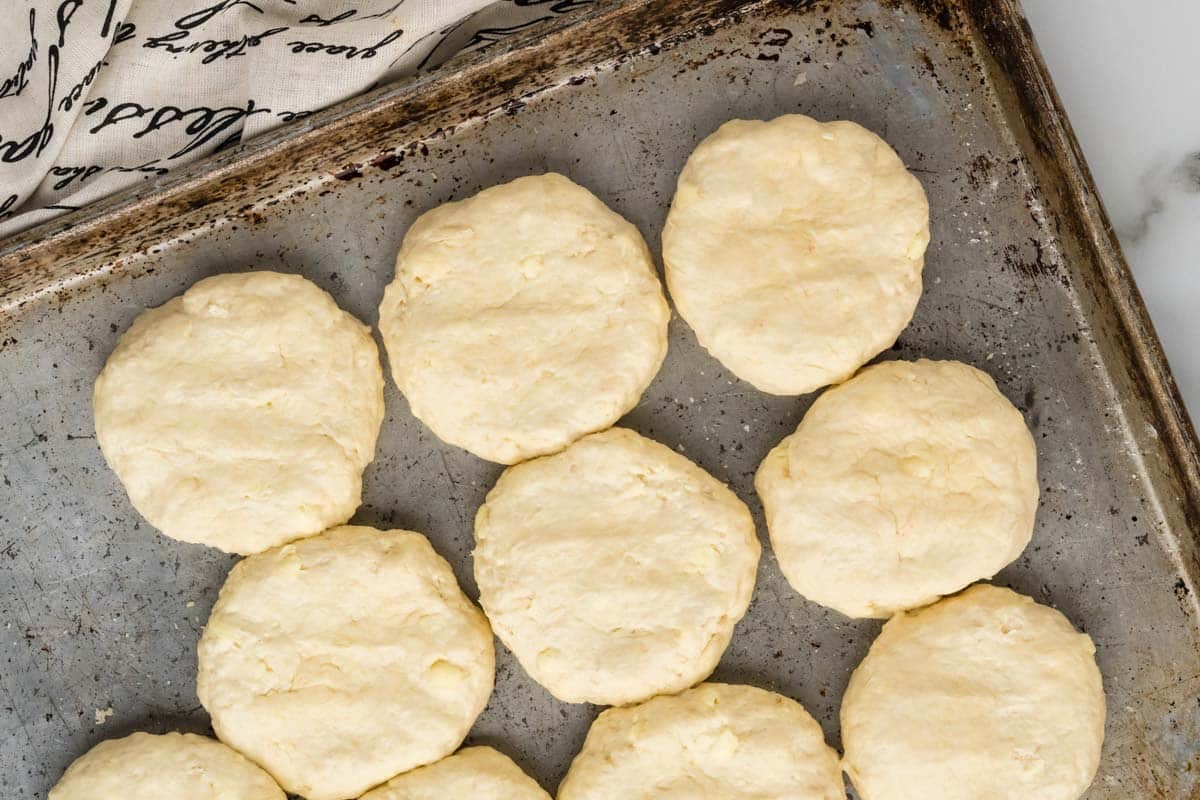 A baking tray with uncooked biscuit dough is arranged on a metal surface, ready to be baked. A patterned cloth is partially visible in the top left corner.