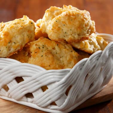 A white woven basket filled with golden-brown, herb-infused biscuits sits on a wooden board.