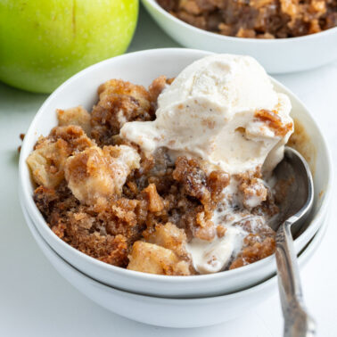 A bowl of apple crisp topped with a scoop of vanilla ice cream, served with a spoon. Two green apples are in the background.