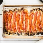 A baking sheet lined with foil holds several cooked strips of bacon, a classic choice for budget-friendly family meals. A cup of coffee, tongs, fork, knife, and a napkin are on the white table nearby.