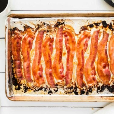 A baking sheet lined with foil holds several cooked strips of bacon, a classic choice for budget-friendly family meals. A cup of coffee, tongs, fork, knife, and a napkin are on the white table nearby.