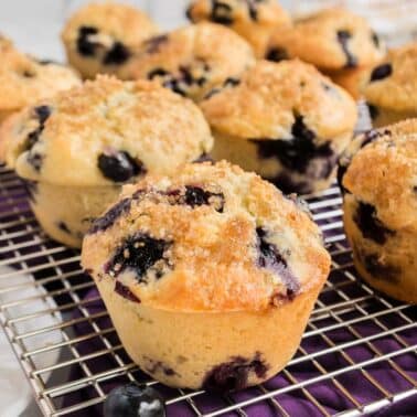 Freshly baked muffins with blueberries and a crumbly topping are cooling on a silver wire rack, placed on a purple cloth.