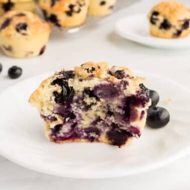 An old-fashioned blueberry muffin on a white plate with whole blueberries around. More muffins are visible in the background.