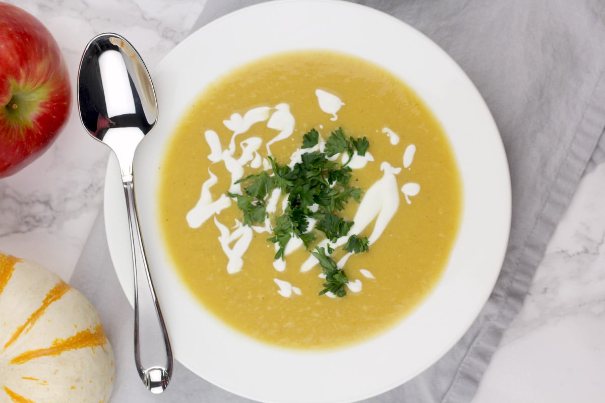 A bowl of pumpkin soup garnished with cream and parsley is placed on a gray cloth, accompanied by a spoon, a red apple, and a small striped gourd.