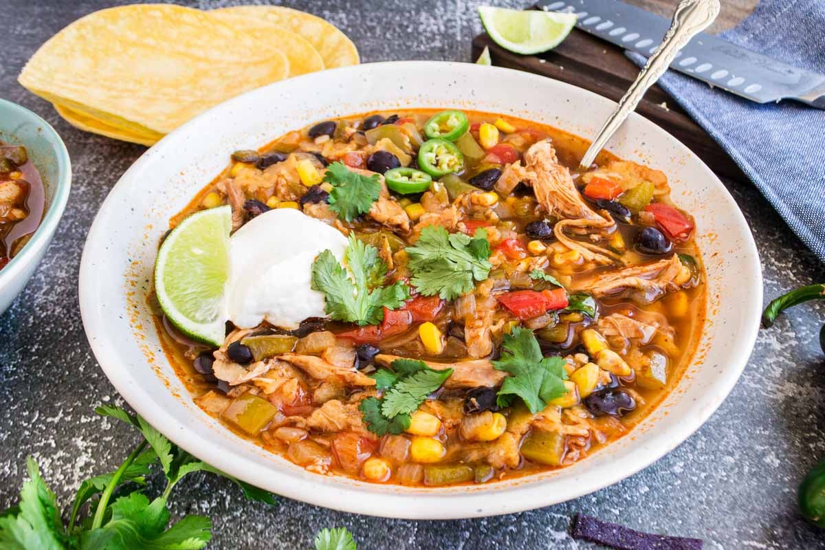 A bowl of chicken tortilla soup garnished with cilantro, sliced jalapeños, sour cream, and a lime wedge, with corn tortillas on the side. The soup contains beans, corn, and various vegetables.