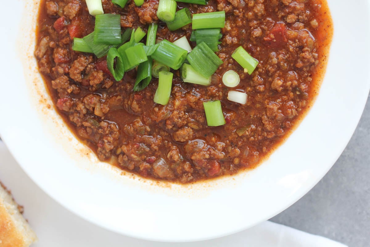 A white bowl of chili topped with chopped green onions, placed on a gray surface with a bread roll partially visible on the side.