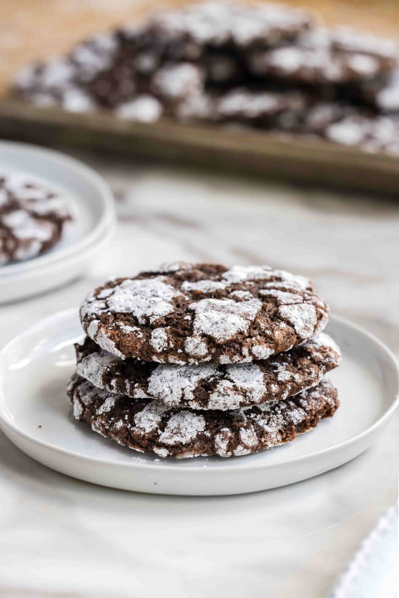 Three chocolate crinkle cookies stacked on a white plate, with more cookies on a baking sheet in the background.