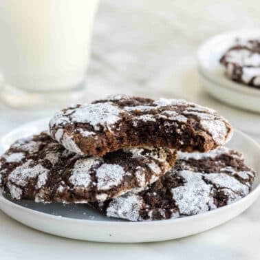 A plate of three chocolate brownie crinkle cookies with powdered sugar on top; one cookie has a bite taken out of it. A glass of milk is in the background.