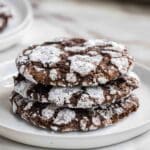 Three chocolate crinkle cookies dusted with powdered sugar stacked on a white plate.