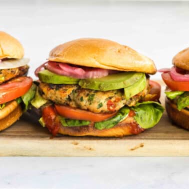 Three jalapeno turkey burgers with lettuce, tomato, avocado, pickled onions, and a toasted bun on a wooden board.