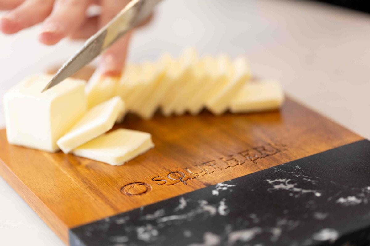 A person slices a block of butter on a wooden cutting board with the word "Squisito" engraved on it.