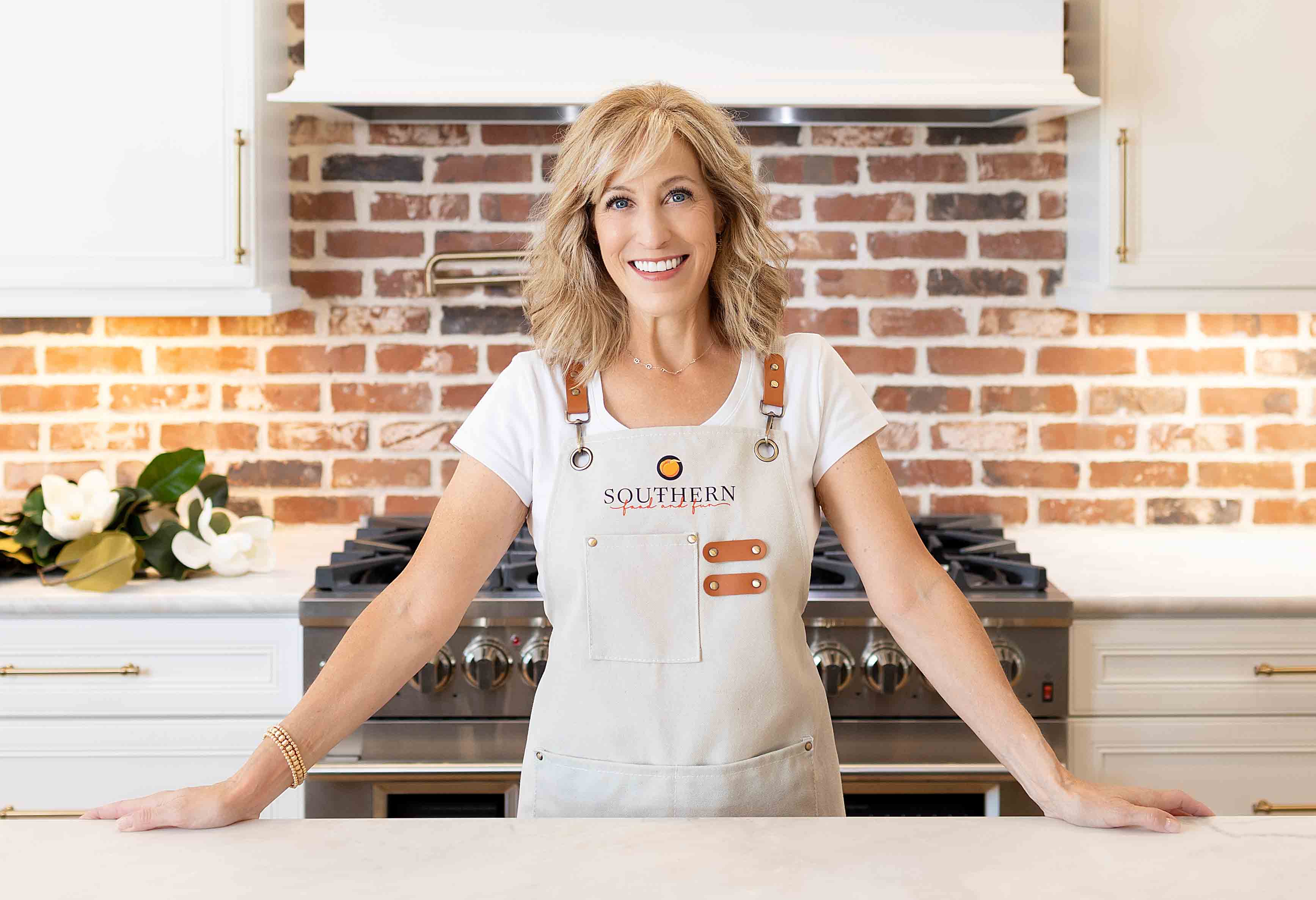 A woman with blond hair, wearing a white shirt and apron, stands smiling in a kitchen with a brick backsplash and stainless steel stove.